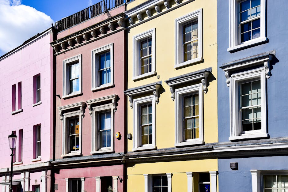 Exterior view of a colourful residential building in Notting Hill with multiple bay windows framed by ornate white trims, painted in pastel shades of pink, yellow, blue, and lilac. The building featur