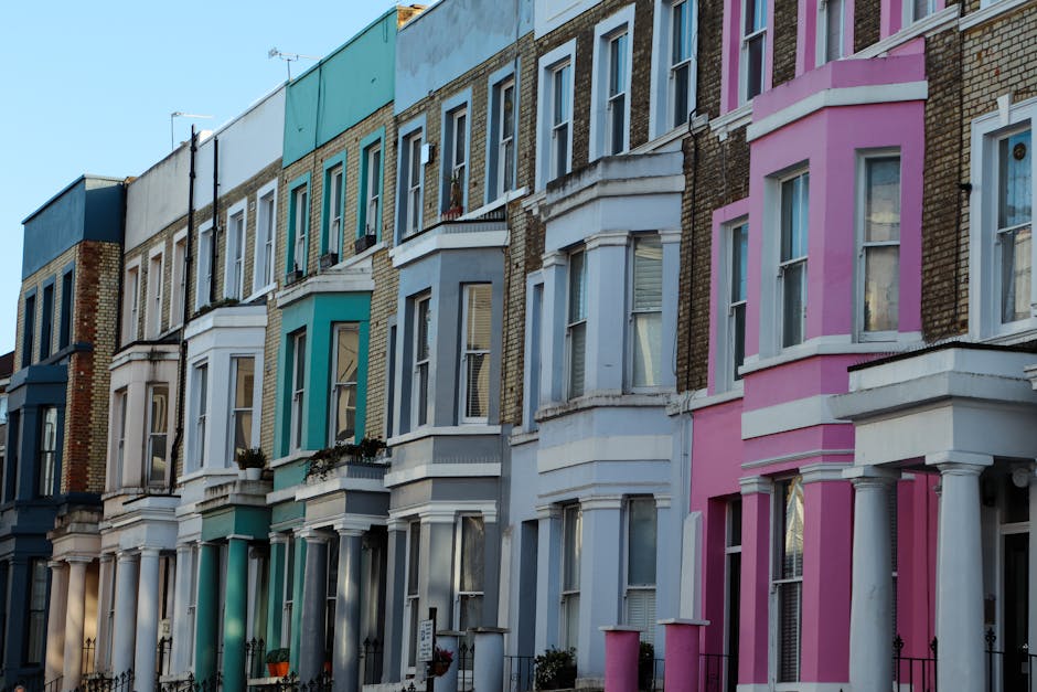 A busy street scene in Notting Hill during daylight with pedestrians walking across the road and along the pavement. The street is lined with multi-storey buildings housing various shops, restaurants,