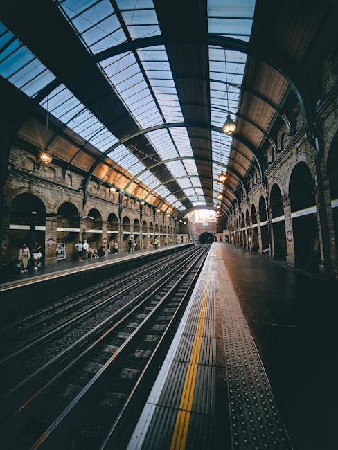 The image depicts a spacious underground railway station with a high, arched glass and metal ceiling allowing natural light to illuminate the platform area. The station features brick walls with multi