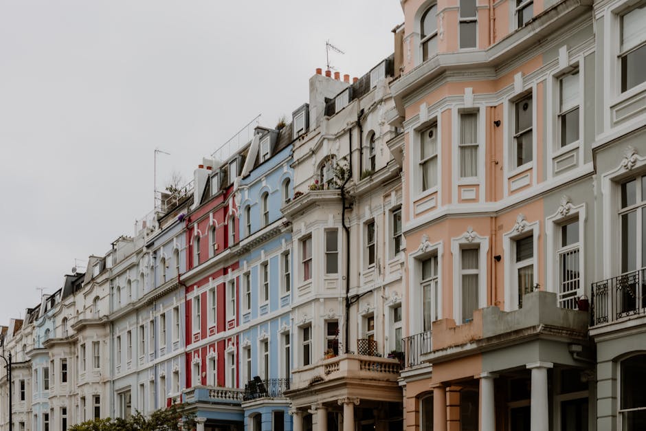 Photograph of a row of residential buildings in Notting Hill, featuring Victorian-style facades painted in pastel blue, with decorative white trim and ornate window frames. The building on the right i