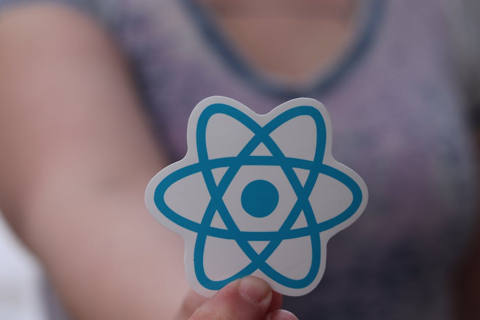 A person holding a small, flat sticker featuring a blue atom symbol with a central dot and intersecting elliptical orbits, in front of a blurred background of an indoor space. The sticker is being hel