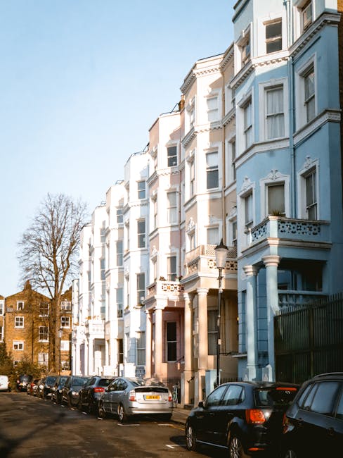A row of multi-storey terraced houses in Notting Hill with distinct blue, beige, and green facades, each featuring large sash windows, decorative molding, and bay windows. The buildings are made of br